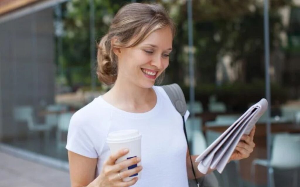 Nasmejana devojka, nosi u ruci kafu za poneti i novine