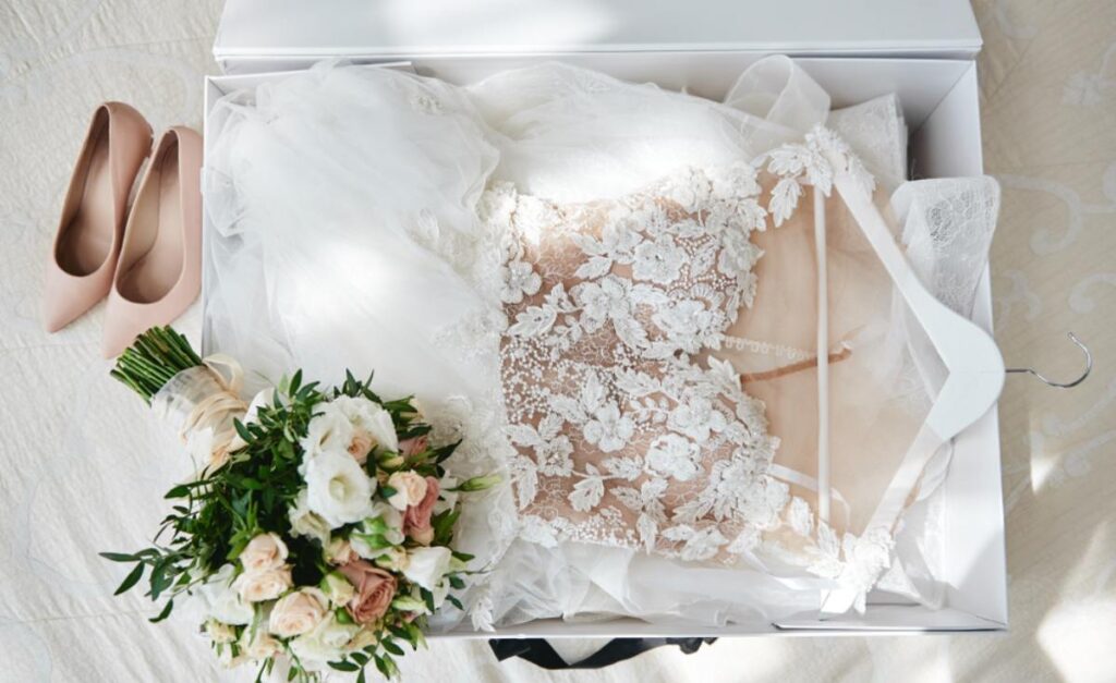 Na slici je prelepa venčanica u kutiji, pored leži bidermajer, a u pozadini se vide svečane cipele.
