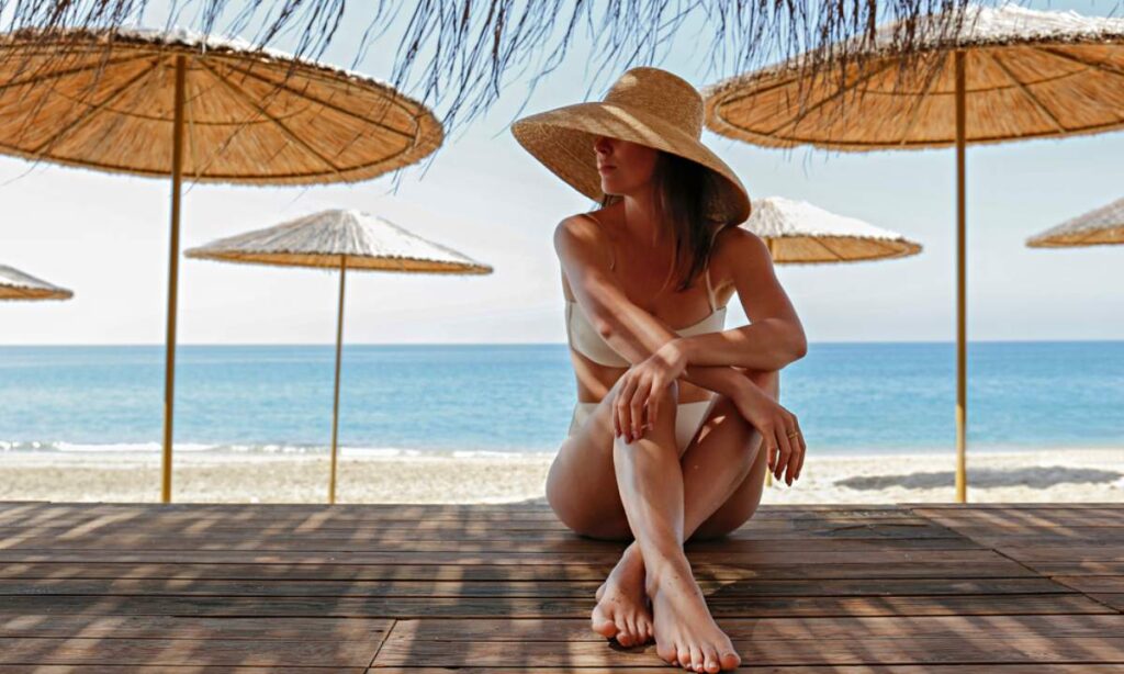 Žena na plaži, sedi u hladovini sa šeširom širokog oboda.
