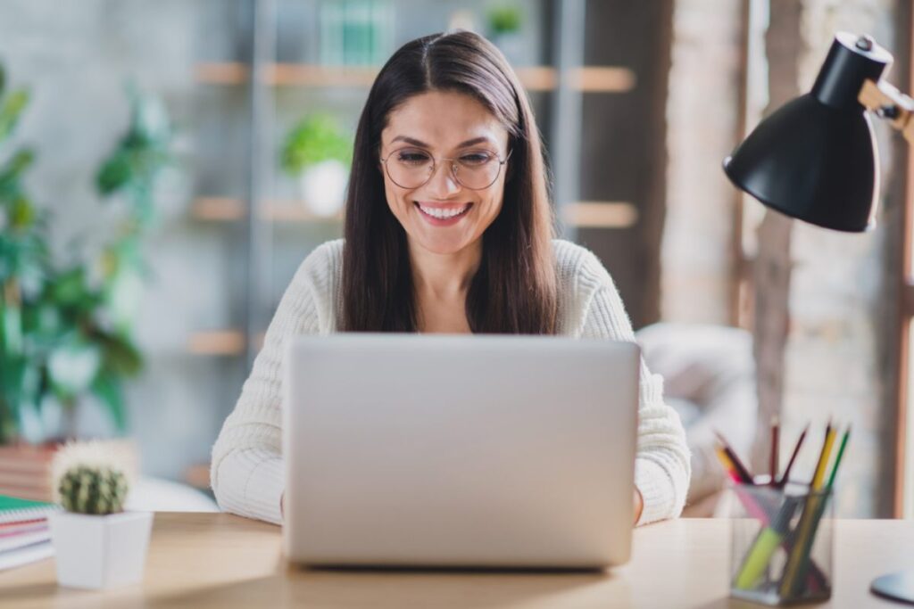 Žena sa naočarima i osmehom na licu rdi na kompjuteru.
