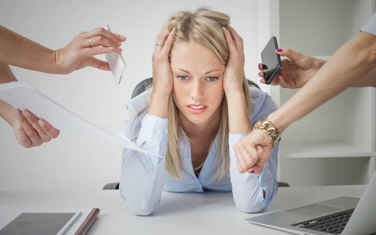 devojka u kancelariji pod stresom, drži se za glavu, dok joj gomila ruku sa strane nudi dokumenta, mobilni telefon itd.