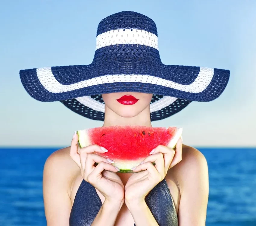 Žena sa šeširom velikog oboda sa obe ruke drži krišku lubenice. U pozadini se vidi more.