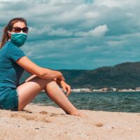 zena sedi u pesku na plazi sa zastitnom maskom