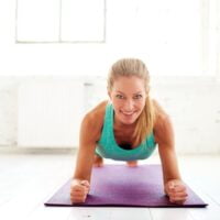 Nasmejana žena u tirkiznom dresu radi plank izdržaj na ljubičastoj prostirci, u sali za vežbe.