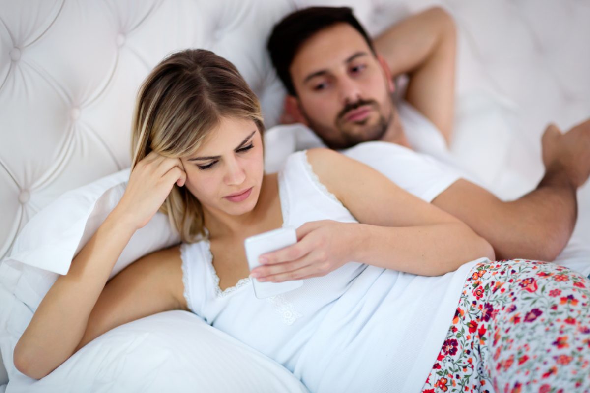 Žena gleda u mobilni telefon, njen partner u krevetu gleda preko njenog ramena s kim se dopisuje