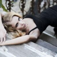 Lepa plavuša leži na stepenicama u crnoj haljini sa bretelama, sa rukama podignutim iznad glave.