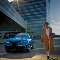 Alfa Romeo - Tonale parkiran ispred moderne zgrade, ispred korača lepa žena u kaputu boje kamilhara.