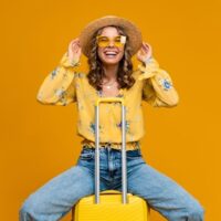 Devojka sedi na koferu, na glavi joj je slamnati šešir. Nosi farmerke i žuru bluzu sa cvetovima.