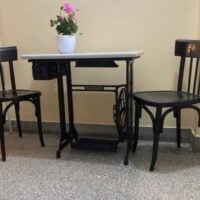 Stara Singer mašina pretvorena u sto. Kutak je napravljen dodatkom dve crne stolice, a na stolu je saksija sa cvećem ružičaste boje.