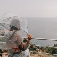 Devojka sa providnim kišobranom sedi na klupi i gleda more po kome pada kiša