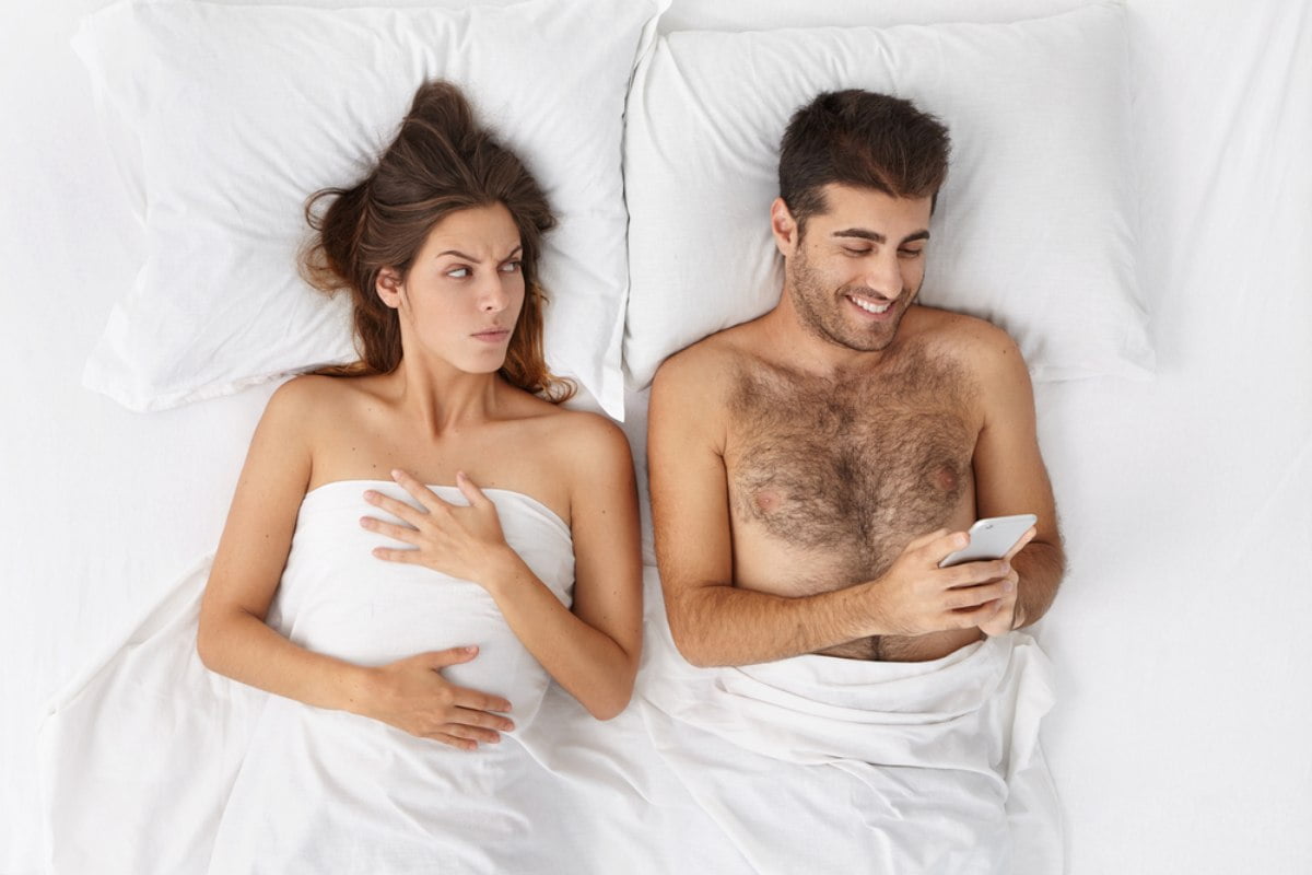 Mladi par u krevetu, devojka popreko gleda mladića koji zaneto gleda u mobilni
