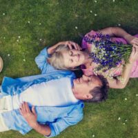 Par leži na travnjaku. Žena drži buket cveća, partner je mazi po glavi.
