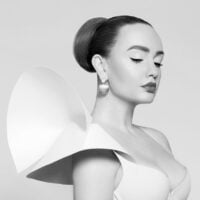 Žena-manekenka, koja malte ne izgleda kao skulptura. Ima obnažena ramena, a haljina koju nosi ima neobične ogrome kratke rukave, kao epolete, krilca. Dekolte je izrezan.