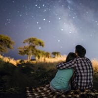 Zagrljeni par koji sedi u polju, vide se sa leđa i posmatraju zvezdano nebo.