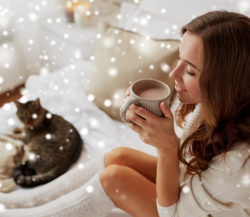 Žena sedi na sofi i uživa pijući toplu čokoladu, u pozadini se vidi mačka. Napravljen je efekat kao da promiču pahulje.