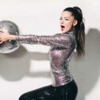 Smeša devojka sa visoko podignutom kosom, drži disko kuglu obučena u bluzu od sjanog materijala i gliter pantalone