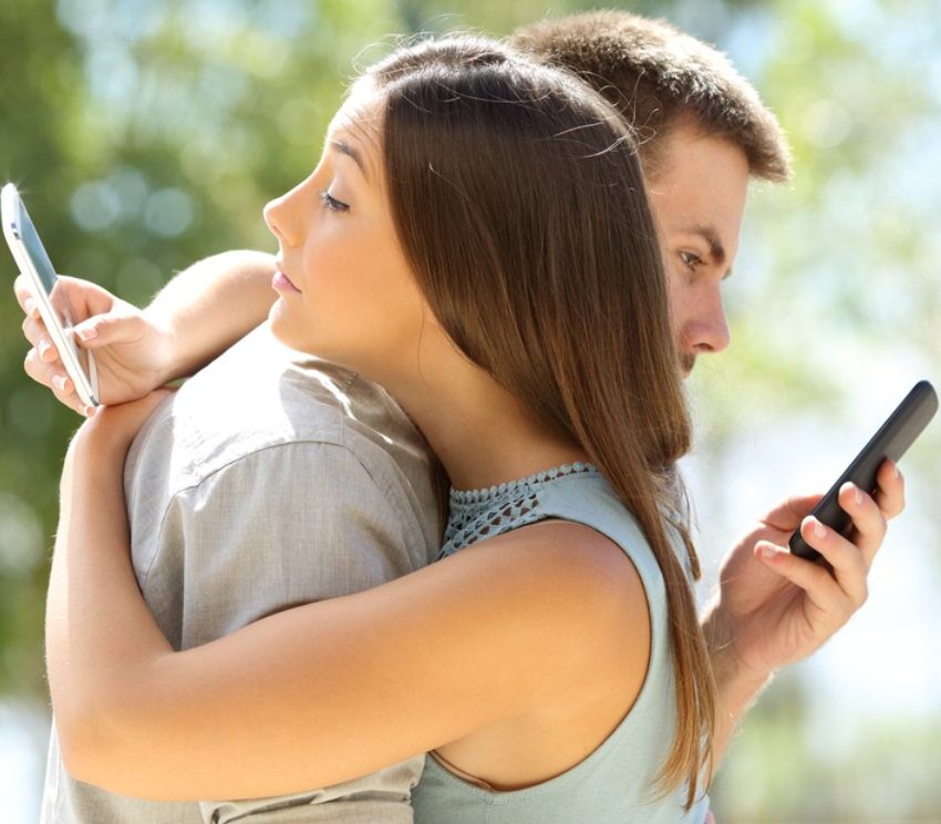 Muškarac i žena u zsagrljaju. Žena preko ramena partnera gleda u mobilni telefon, dok muškarac preko ženinog ramena gleda us voj mobilni telefon.