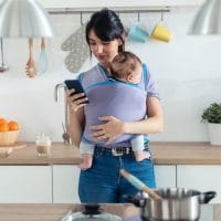 Mama drži bebu u kengur torbi i pretražuje ili nešto gleda na mobilnom telefonu.