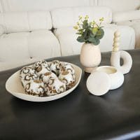 Sto u dnevnoj sobi na kome je činija sa dekorativnim kuglama, svećnja i vaza sa lepim cvetnim aranžmanom od belog cveća.