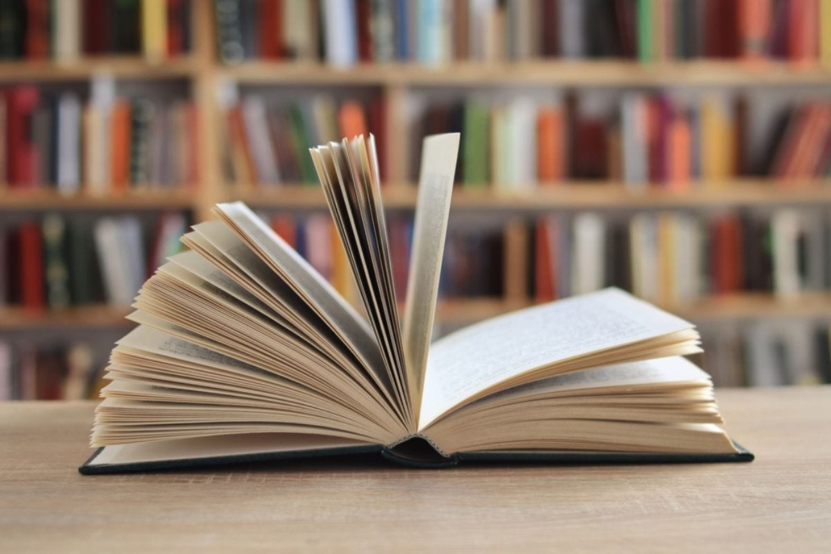 Otvorena knjiga, stranice, a u pozadini e vidi biblioteka sa knjigama.