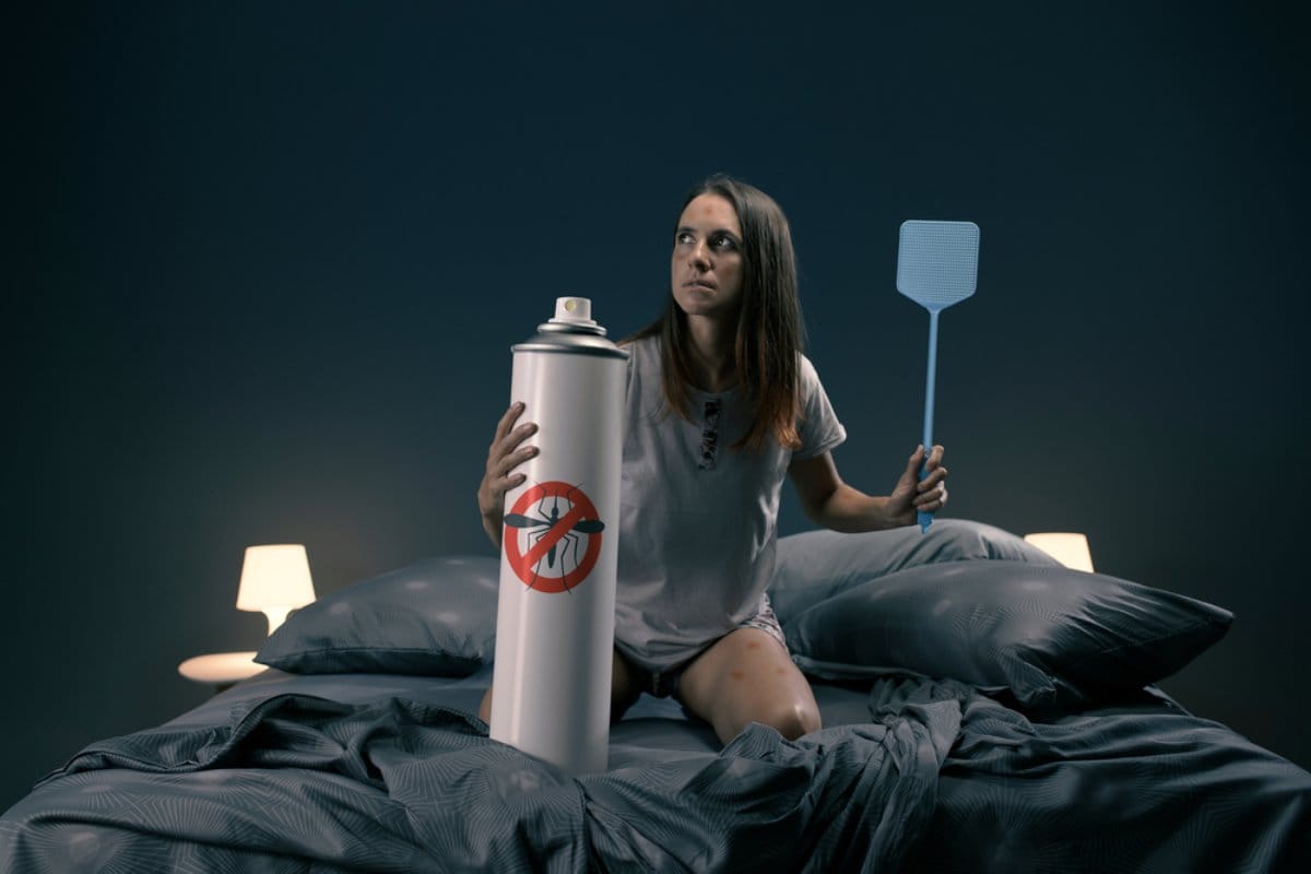 Žena u spavaćoj sobi, izujedali su je komarci, ima palicu za ubijanje komaraca u jednoj ruci, u drugoj drži preuveličani repelent. Ljuti izraz lica.