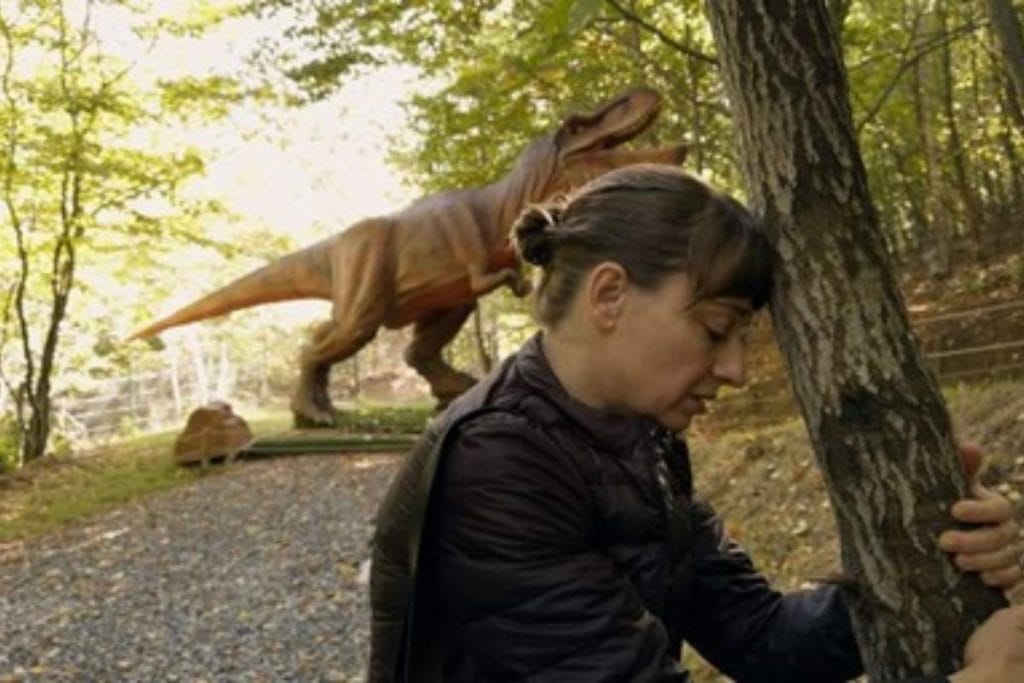 Glumica u šumi, naslonila je glavu na drvo, a u pozadini se vidi dinosaurus.
