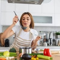 Žena u kuhinji sprema ručak i isprobava ukus, okolo je razno povrće koje koristi u kuvanju.