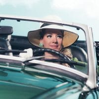 Žena vozi retro sportski auto, ima šešir sa širokim obodom i velike akril rezeda zelene minđuše.
