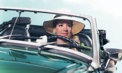 Žena vozi retro sportski auto, ima šešir sa širokim obodom i velike akril rezeda zelene minđuše.
