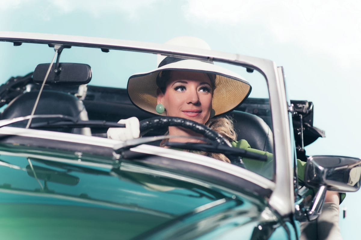 Žena vozi retro sportski auto, ima šešir sa širokim obodom i velike akril rezeda zelene minđuše.
