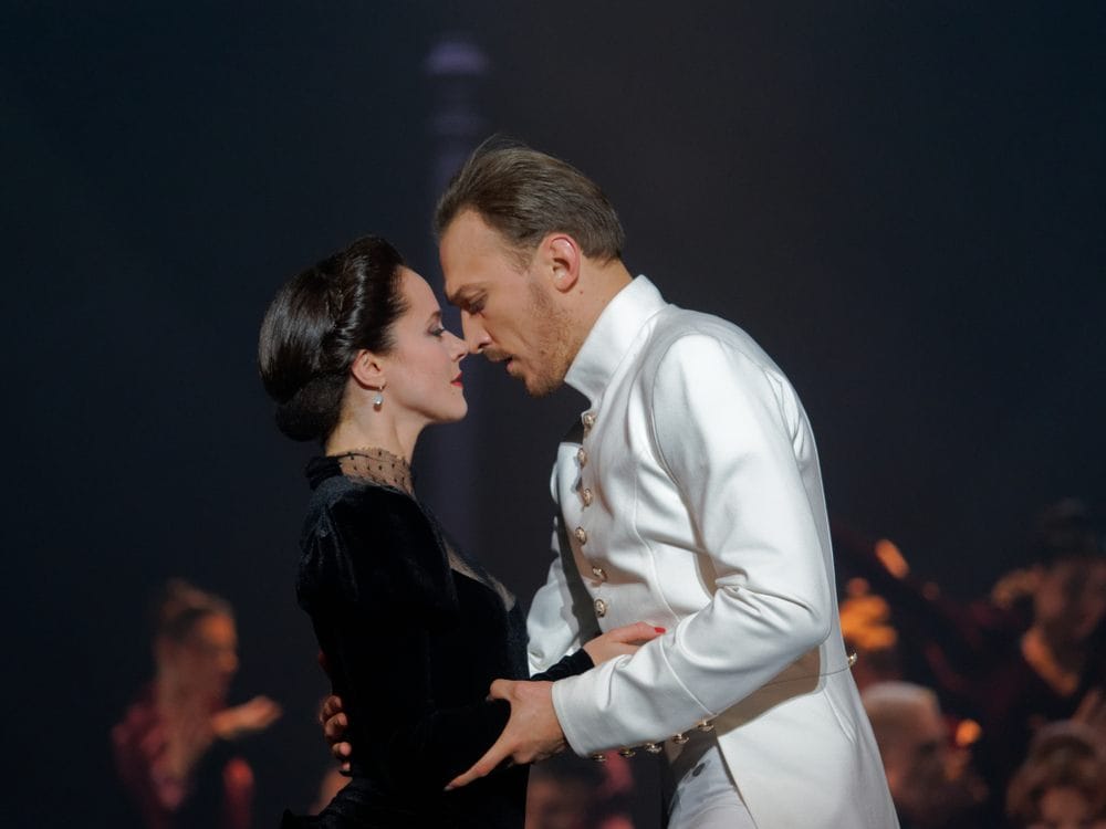 Ana Karenjina, scena iz baletske predstave. Ana je u crnoj somotskoj haljini sa čipkom haljini u zagrljaju oficira Vronskog u beloj uniformi.