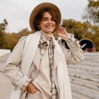 Žena sa bež šeširom, nosi bež rolku, mantil, preko je beli vuneni šal i bež šešir.