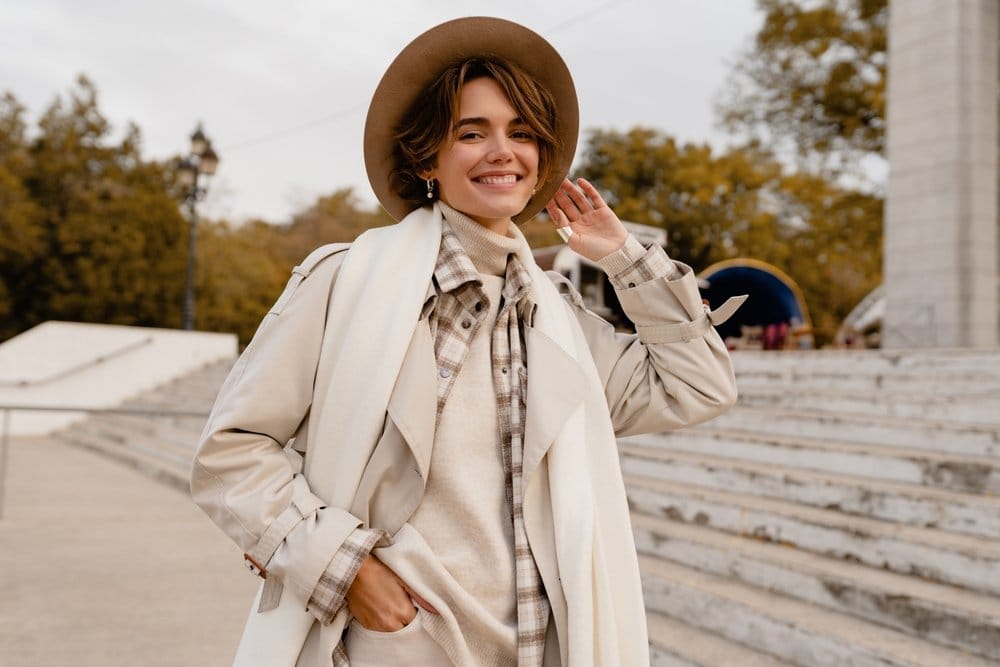 Žena sa bež šeširom, nosi bež rolku, mantil, preko je beli vuneni šal i bež šešir.