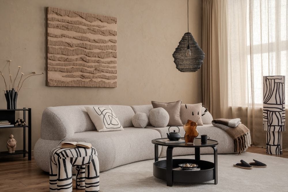 Dnevna soba, bež sofa sa puno jastučića, moderna slika na zidu, neobično zebrasto kanabe, moderni stočić i lampa. Tepih na kome su kućne papuče,