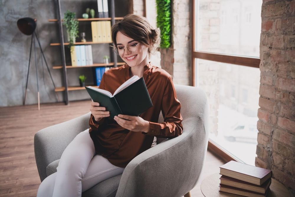 Žena koja sedi u fotelji i sa uživanjem čita knjigu,