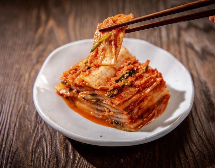 Kim chi, naslagani listovi kimči kupusa u tanjiru, preko listova je pasta od čilij. Vide se štapići kojima osoba hvata listiće.