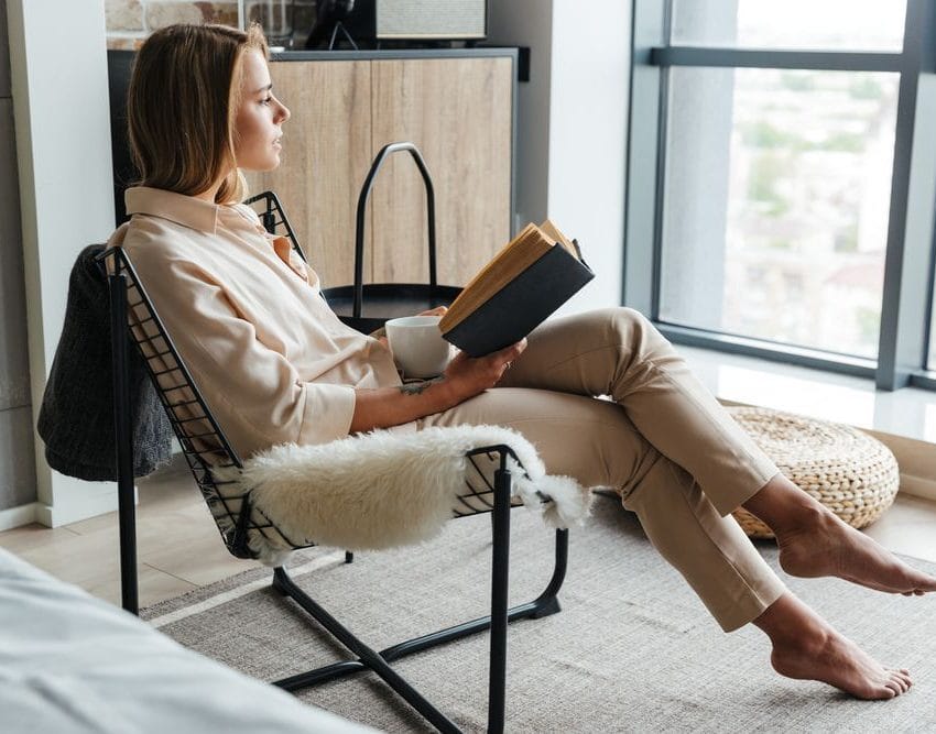 Žena drži knjigu, pogledala je kroz prozor. Tu je i šolja sa čajem.Opuštena je, uživa.