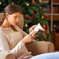 Žena koja sedi u udobnoj fotelji i čita knjigu, u pozadini se vidi okićena jellka.