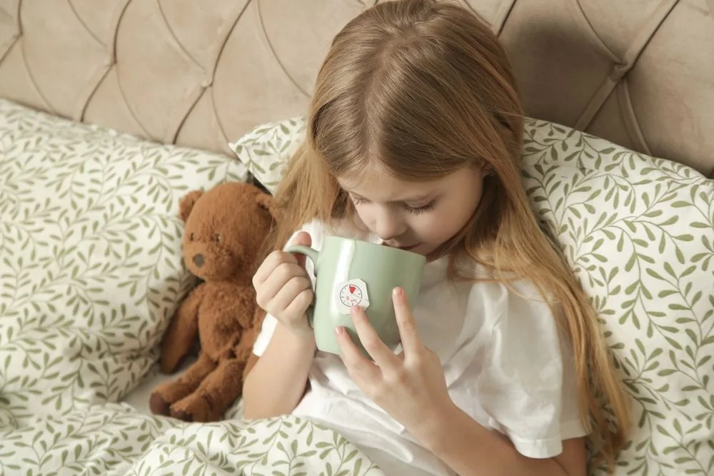 Devojčica u poluležećem položaju, drži medu pored sebe i pije čaj.