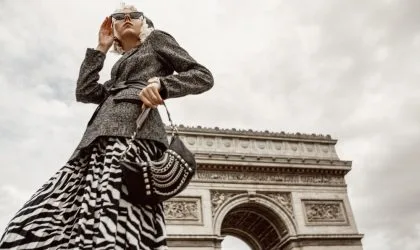 Manekenka u maksi crno-beloj suknji, sivi sako sa kaišpem, naočare za sunce i moderna crna tašna. U pozadini se vidi Trijumfalna kapija u Parizu.