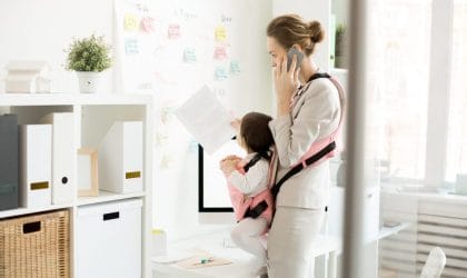 Žena koja radi, gleda u papire i telefonira, u nosiljci koju nosi je beba koja isto gleda u papire. Situacija deluje kontrolisano.