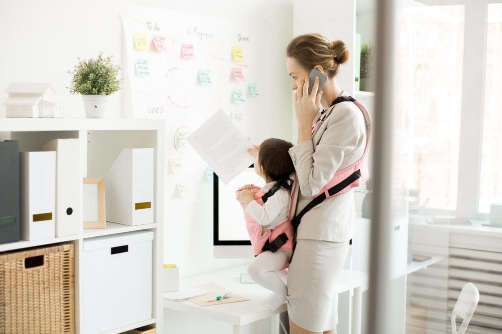 Žena koja radi, gleda u papire i telefonira, u nosiljci koju nosi je beba koja isto gleda u papire. Situacija deluje kontrolisano.