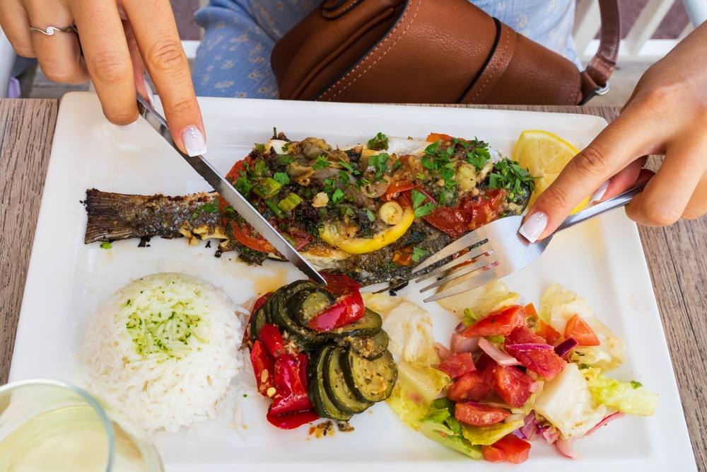 Žena koja u tanjiru ima ribu, uživa u obroku, dodala je i grilovano povrće i salatu.