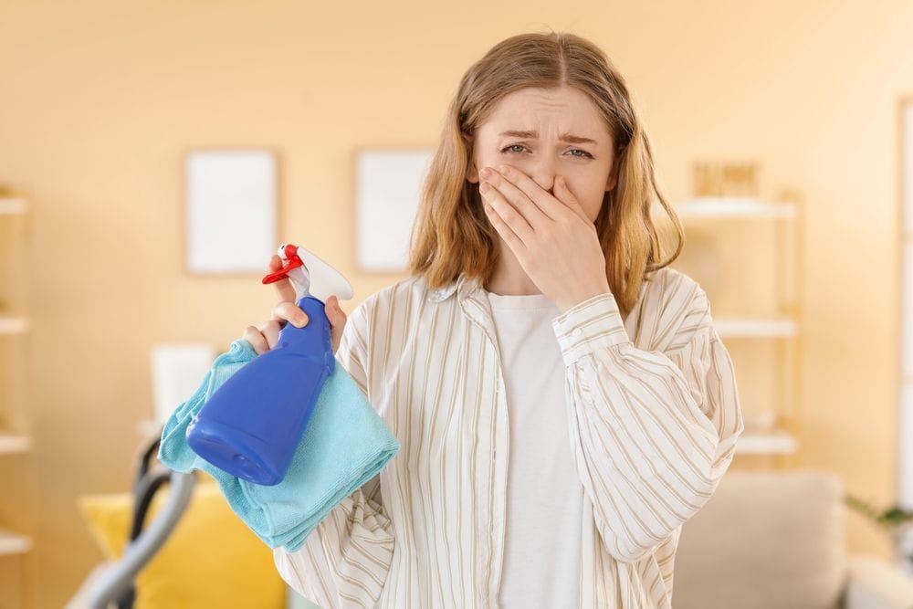 Žena koja drži u ruci sredstvo za či&scaron;ćenje i krpu, a drugom ruku je stavila maramicu na nos.