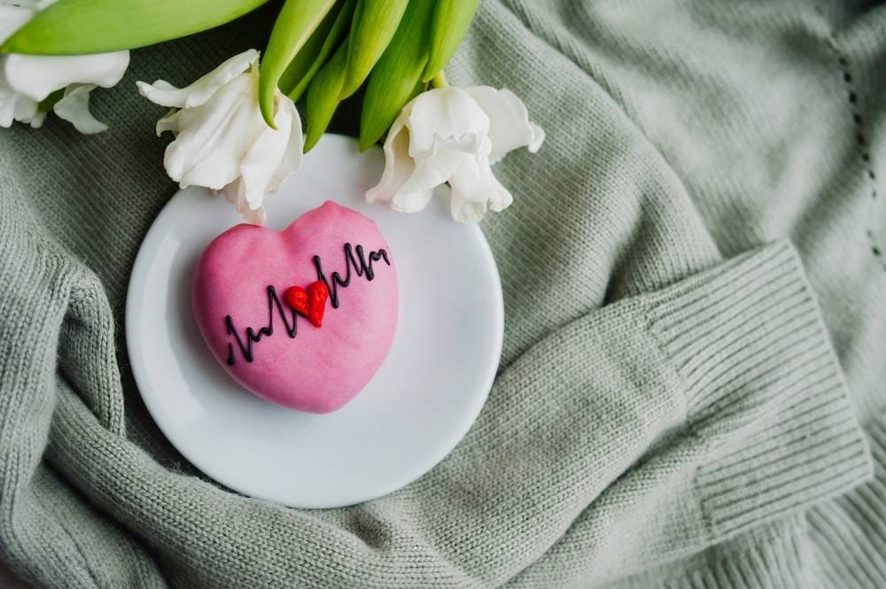 Kolačić u obliku srca na kome je ilustracija EKG-a u pozadini džemper i cveće.