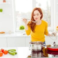 Žena stoji za šporetom, isprobava jelo koje kuva. Na ploči pored je povrće.