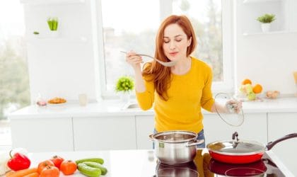 Žena stoji za šporetom, isprobava jelo koje kuva. Na ploči pored je povrće.