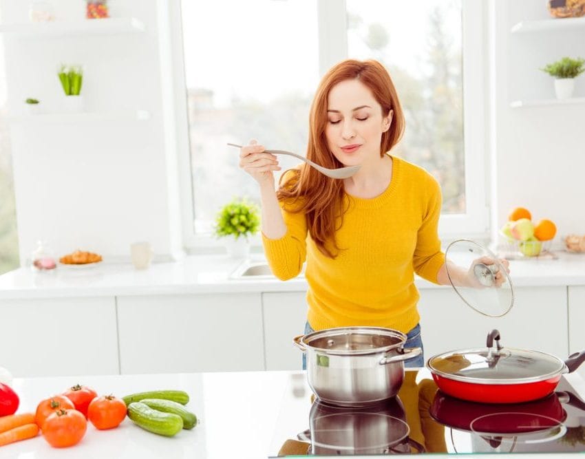 Žena stoji za šporetom, isprobava jelo koje kuva. Na ploči pored je povrće.