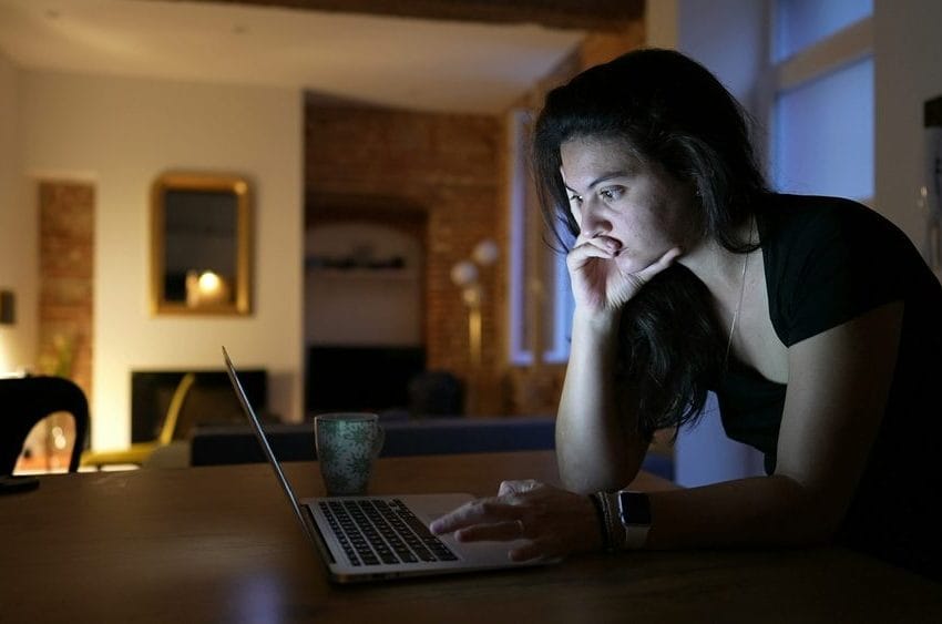 Žena radi na lap top-u u dnevnoj sobi sa slabijim osvetljenjem. Vidi se kako joj lice osvetlajava svetlo lap top-a.