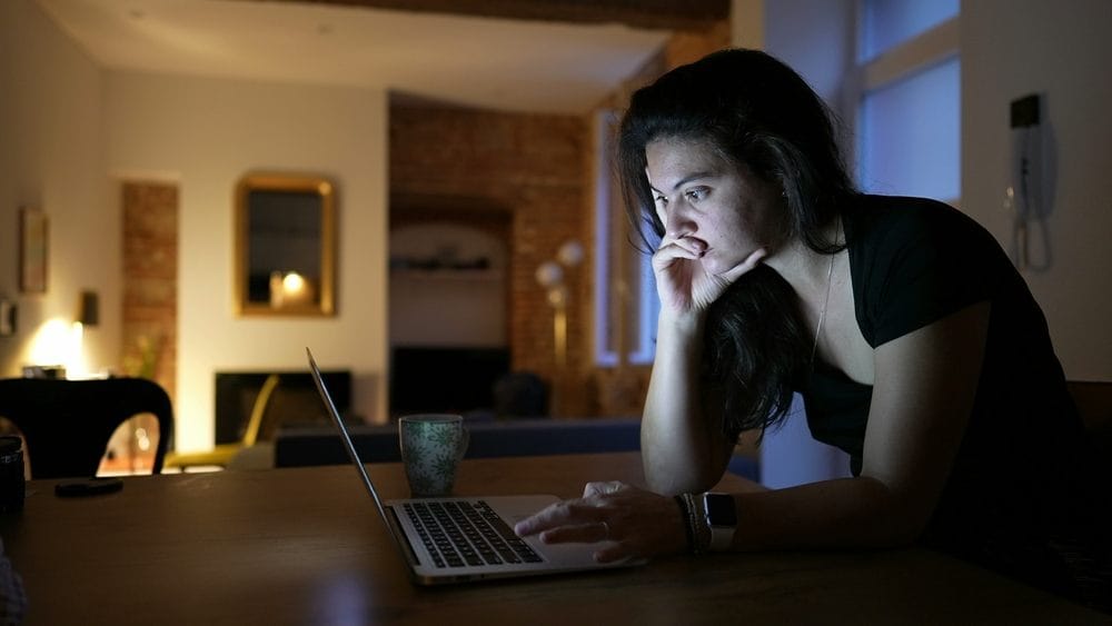 Žena radi na lap top-u u dnevnoj sobi sa slabijim osvetljenjem. Vidi se kako joj lice osvetlajava svetlo lap top-a.